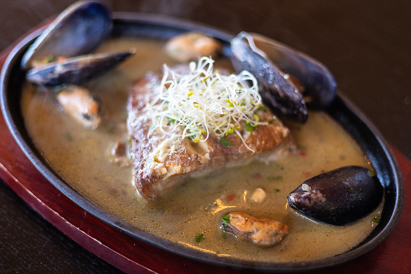 Un filete de pescado sellado con brotes de alfalfa encima, servido en una paila de hierro con choritos (mejillones) y una salsa cremosa. Una excelente muestra de la gastronomía local para quienes buscan dónde comer en Cochamó.